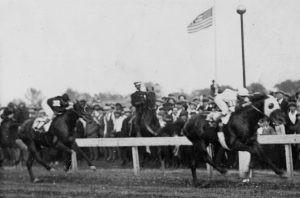 a black and white photo of the winner crossing the finish line in the 1923 Kentucky Derby. there is a crowd watching and an American flag waving in the background.