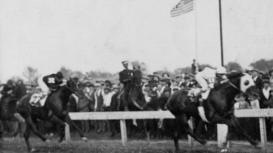 a black and white photo of the winner crossing the finish line in the 1923 Kentucky Derby. there is a crowd watching and an American flag waving in the background.