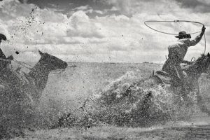 two men on horseback walking in deep water. one man has a rope lassoing above his head